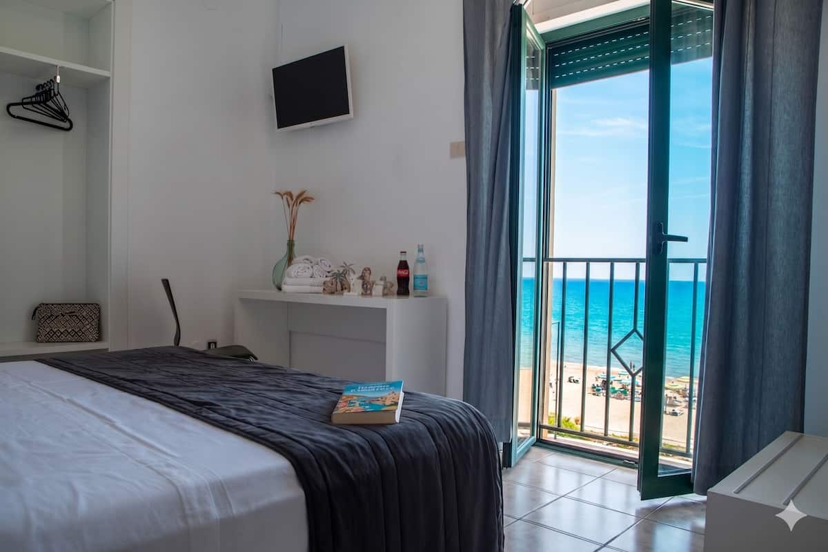 Un dormitorio con cama, una mesita de noche con un libro, televisor montado en la pared y vista a la playa a través de una puerta corrediza.