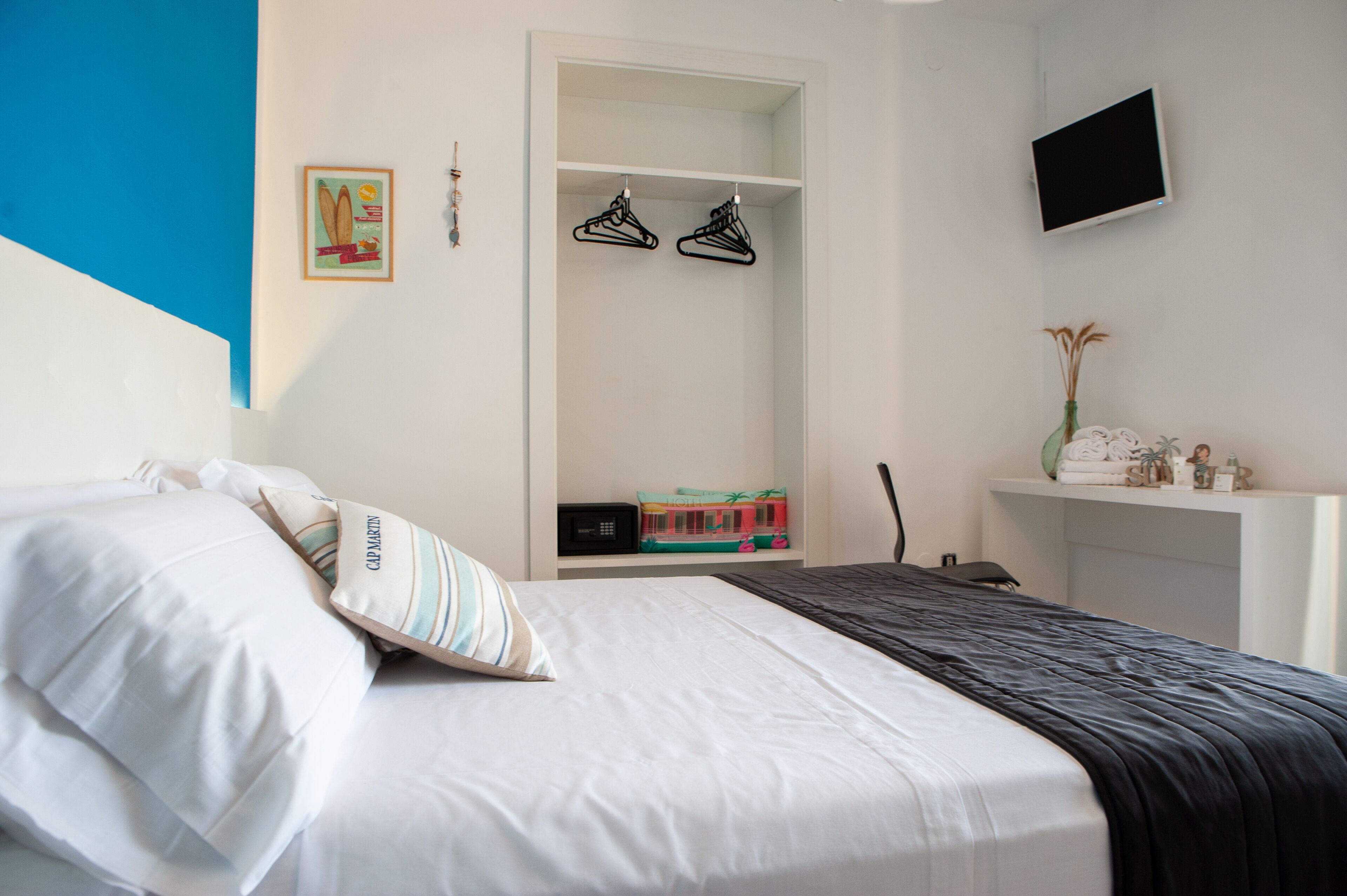 Ein ordentlich bezogenes Bett mit weißen Leinen und gestreiften Kissen, ein Kleiderschrank mit Kleidung, ein kleines Regal mit Pflegeprodukten und ein Flachbildfernseher an der Wand.