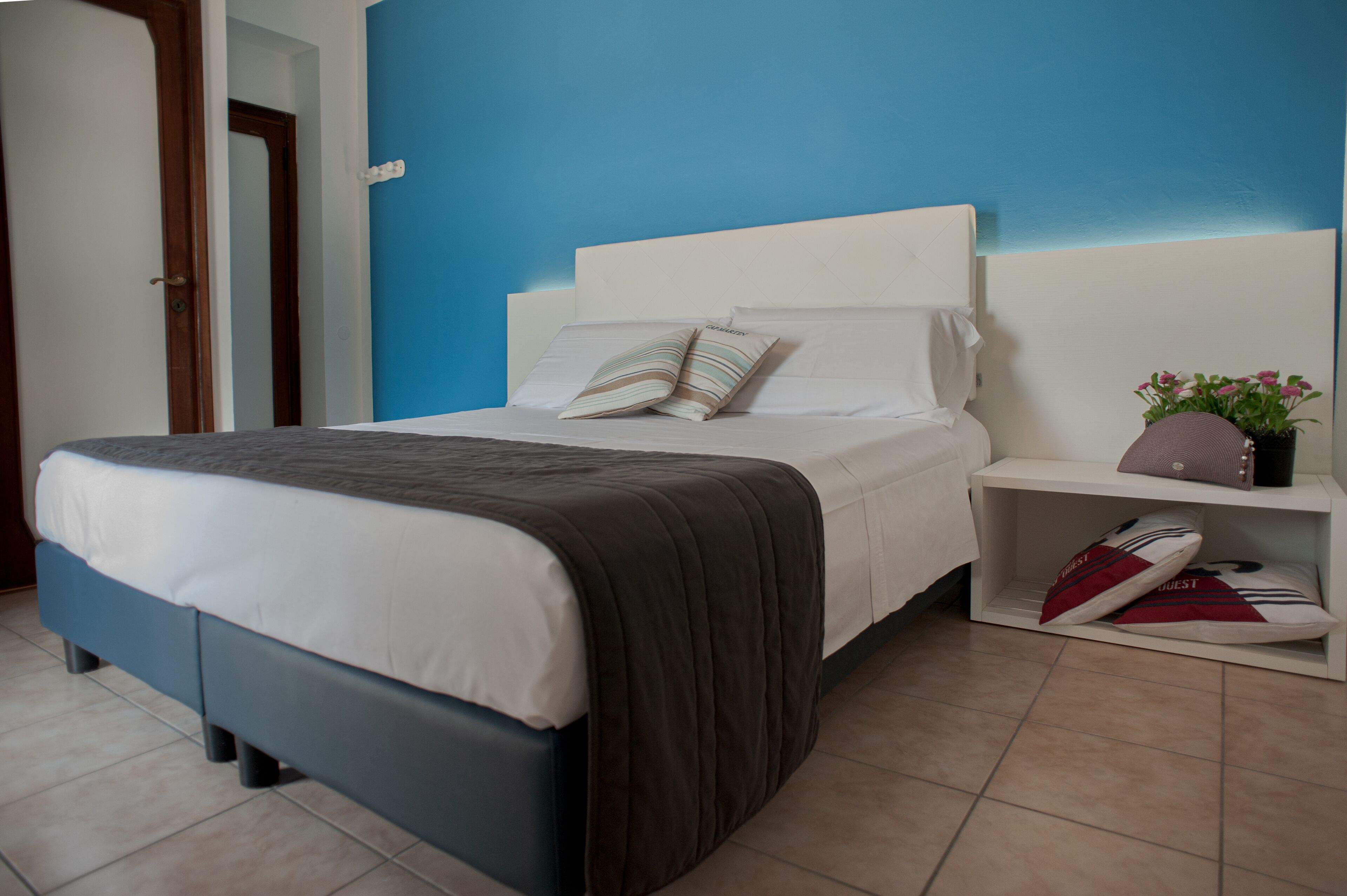 A neatly made bed with a white headboard, a blue wall, and a small bedside table with a plant and slippers.