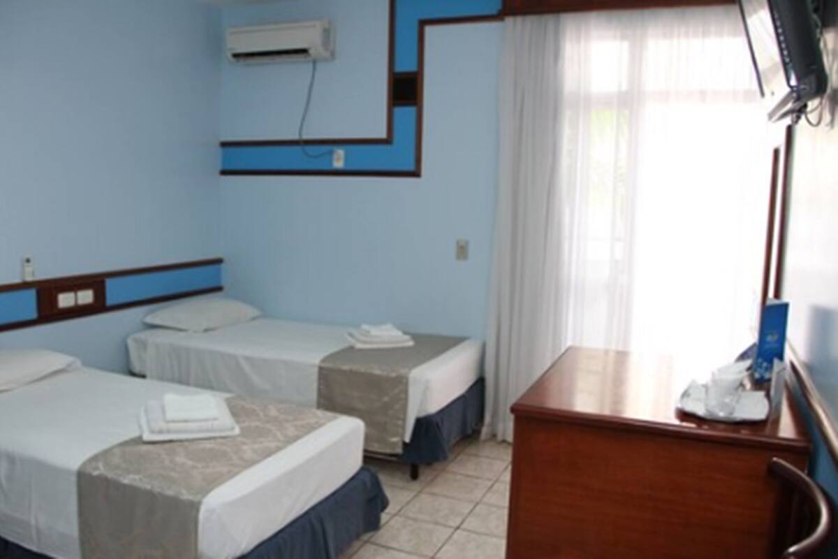 A hotel room with two beds, a desk, a chair, a TV, and an air conditioning unit.