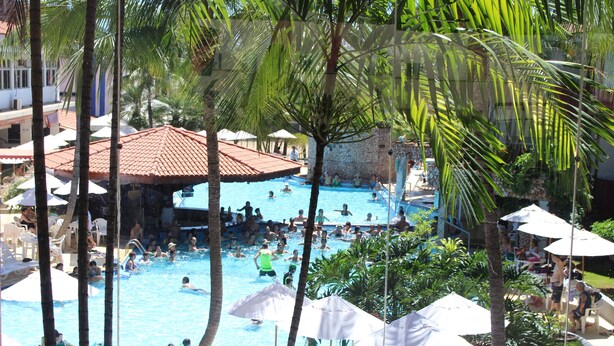 A swimming pool area with multiple people, palm trees, and umbrellas. - Taiyo Thermas Hotel (Caldas Novas)