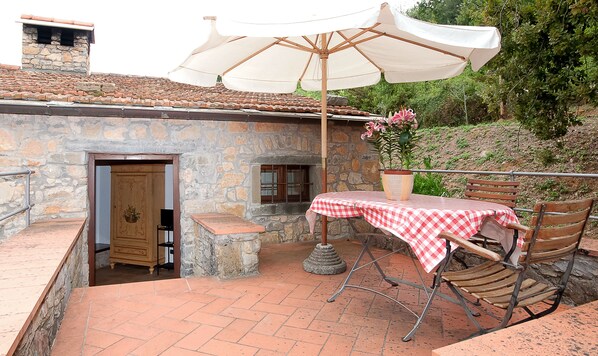 A stone building with a tiled roof, a wooden door, and a small window. There is a table with a checkered tablecloth and a bench under a white umbrella.