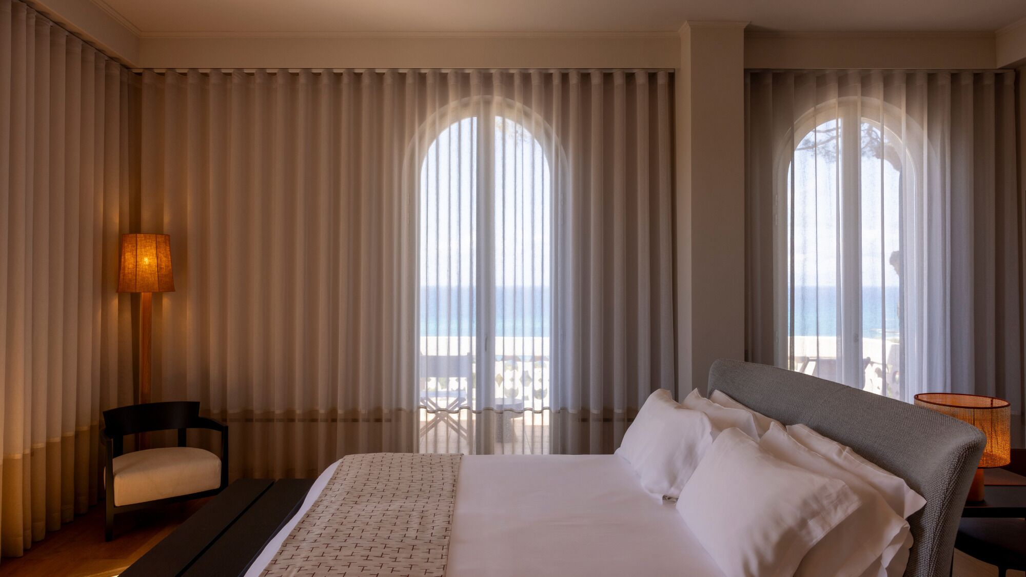 A hotel room with a large bed, a chair, a desk, and a view of the sea through large windows.