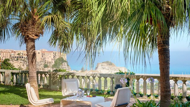 A terrace with two white lounge chairs and a table, overlooking a coastal view with buildings and a clear sky. - Villa Paola (Tropea)