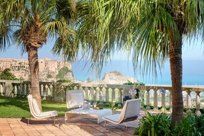 A terrace with two white lounge chairs and a table, overlooking a coastal view with buildings and a clear sky. - Villa Paola (Tropea)