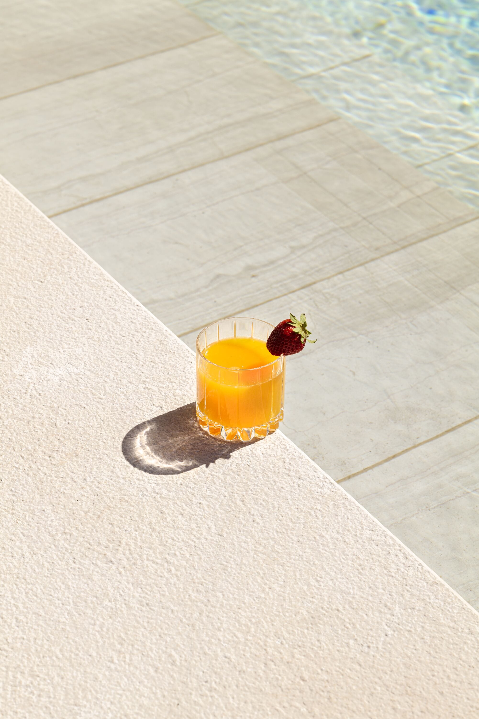 Un vaso de jugo de naranja con una fresa como adorno sobre una superficie rugosa, cerca de una piscina.