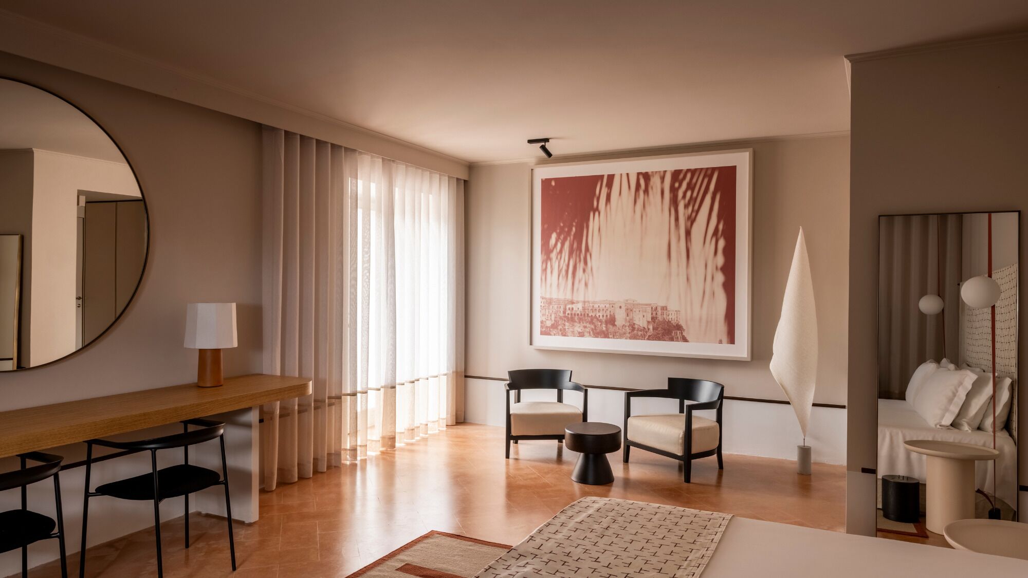 Moderni hotellihuone, jossa on puinen työpöytä, kaksi tuolia, suuri sänky, pyöreä peili ja suuri abstrakti maalaus seinällä.