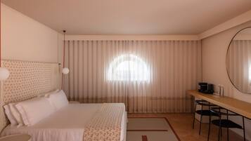 A hotel room with a bed, a desk, a chair, a round mirror, and a window with curtains.