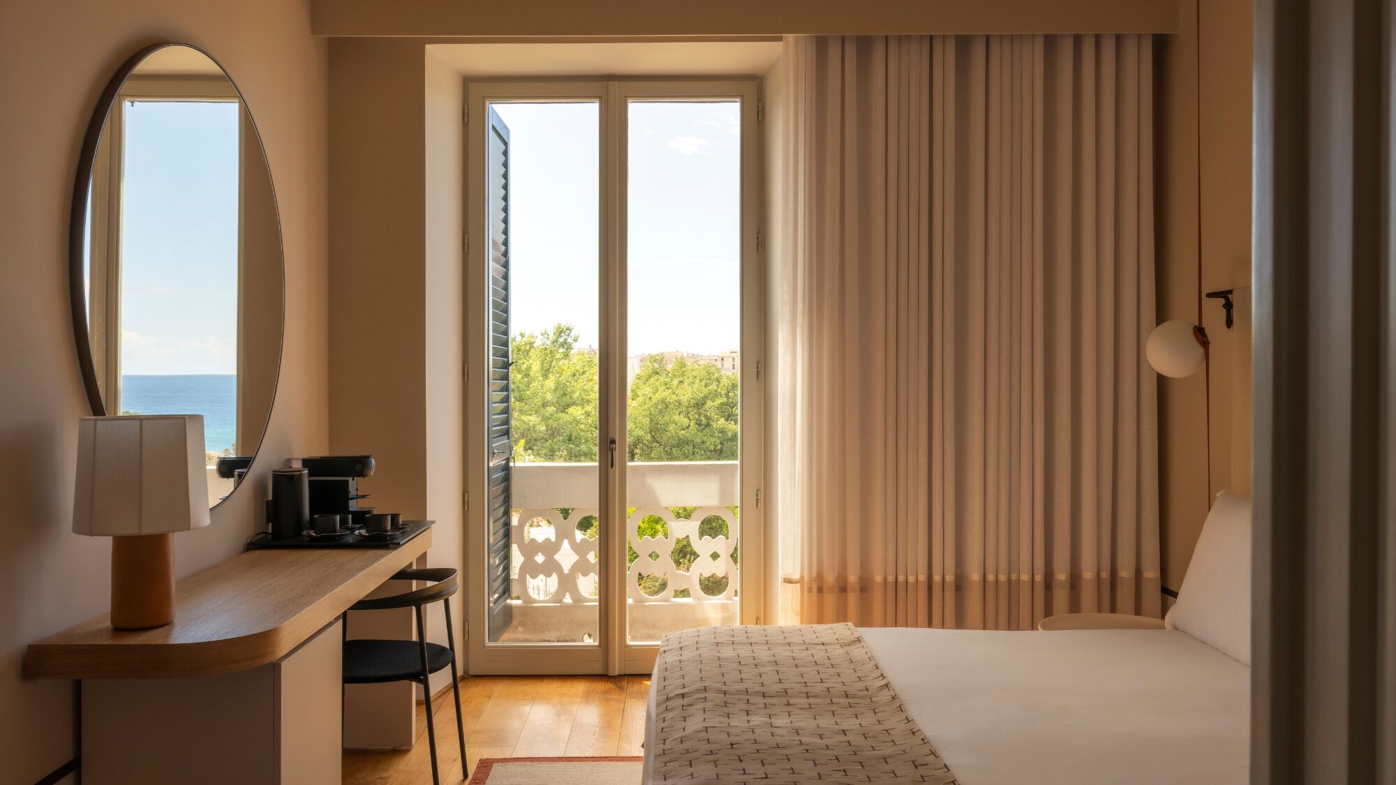 Habitación de hotel con una cama, un escritorio con lámpara, una cafetera y vista al mar a través de una puerta corredera.