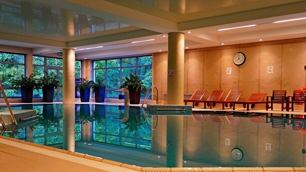 An indoor swimming pool with a clear view of the outside through large windows, a clock on the wall, and several lounge chairs along the poolside.