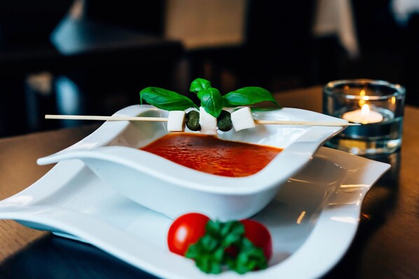 Un plato de cerámica blanca con salsa roja, una brocheta de mozzarella y albahaca, y una vela en vaso sobre una mesa.