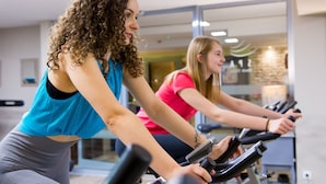 Two individuals on stationary bikes in a gym with mirrored walls and a visible window.