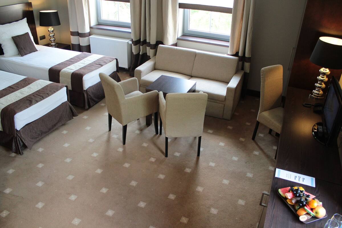 Une chambre d’h ôtel avec deux lits, un canapé, deux fauteuils, une petite table, un bureau avec une lampe et un plateau de fruits.