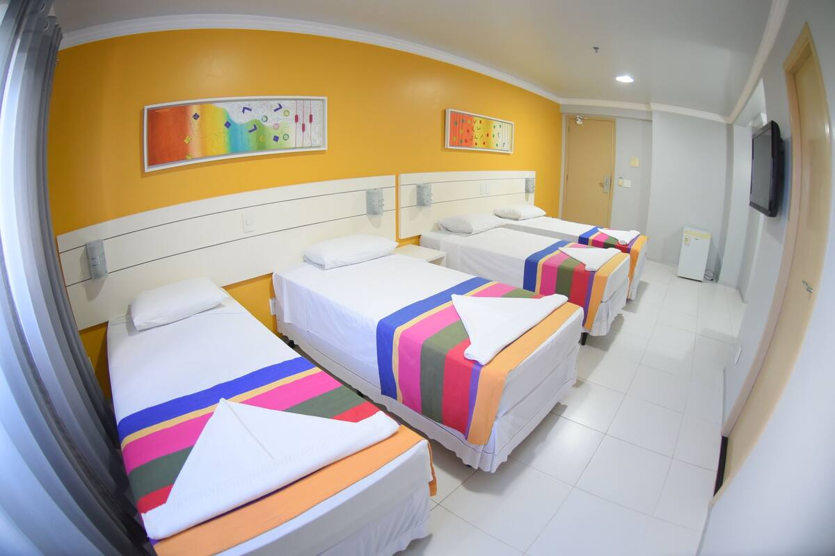 Una habitación de hotel con cuatro camas, cada una con ropa de cama a rayas de colores. Hay dos televisores montados en la pared y un refrigerador en la habitación.