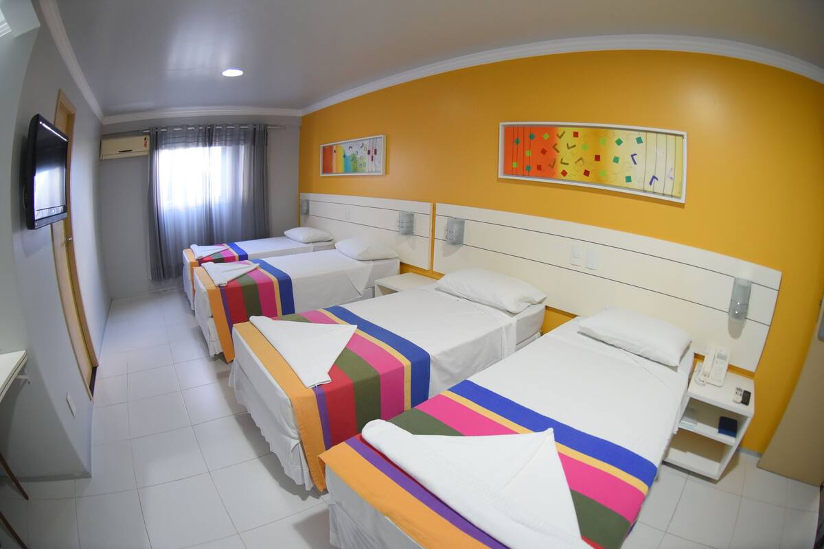 Habitación de hotel con tres camas, cada una con ropa de cama de colores, un televisor de pantalla plana y obras de arte en las paredes.