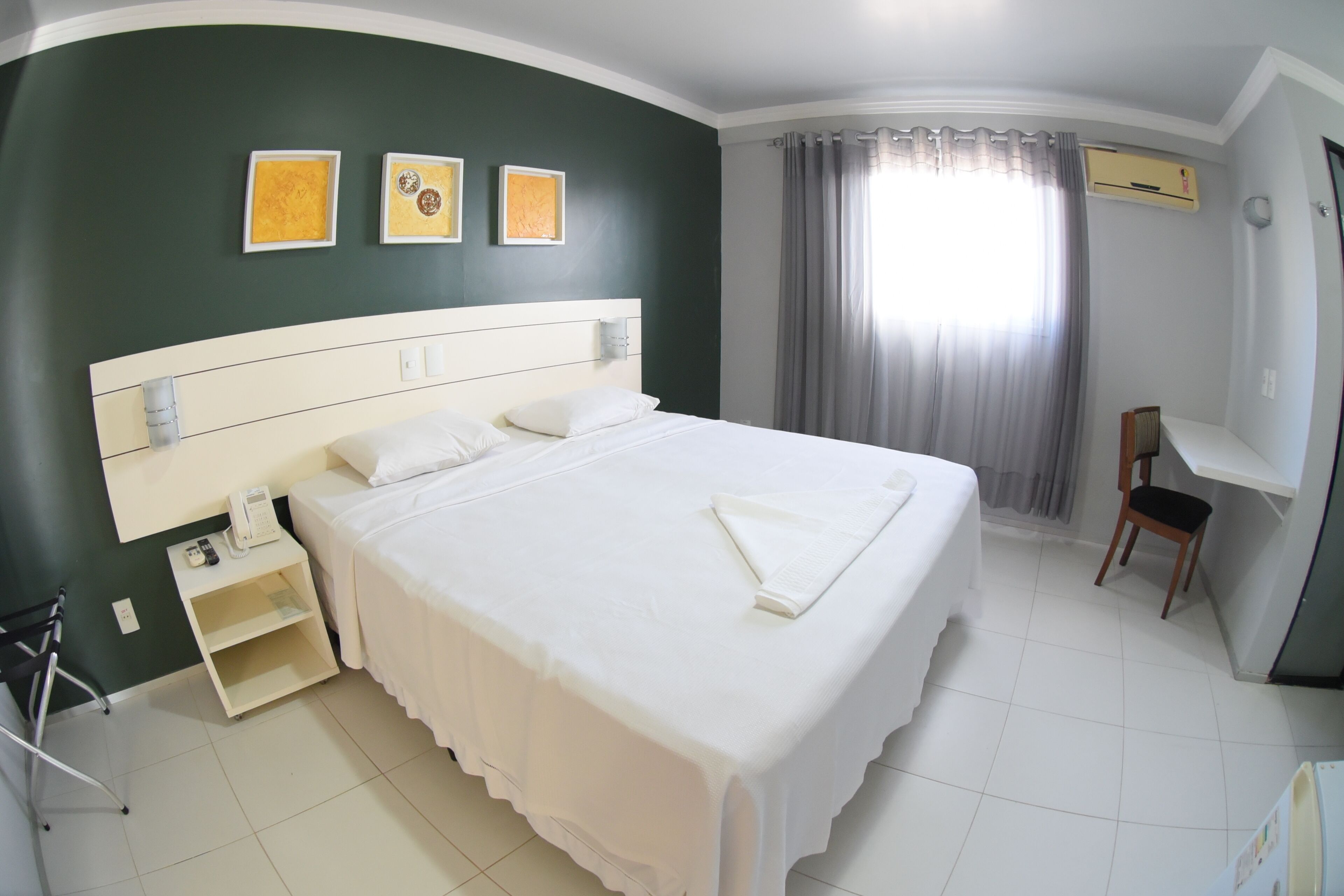 A hotel room with a large bed, a bedside table with a phone, a chair, and a desk. There are three framed pictures on the wall.