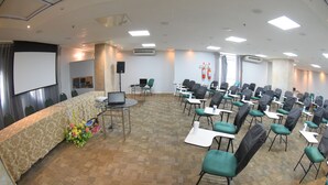Uma sala de conferências com piso carpete, fileiras de cadeiras, uma tela de projetor e uma mesa com um laptop e flores.