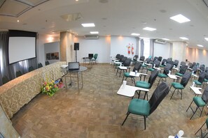 Un salón de conferencias con suelo alfombrado, filas de sillas, una pantalla de proyector y una mesa con una computadora portátil y flores.