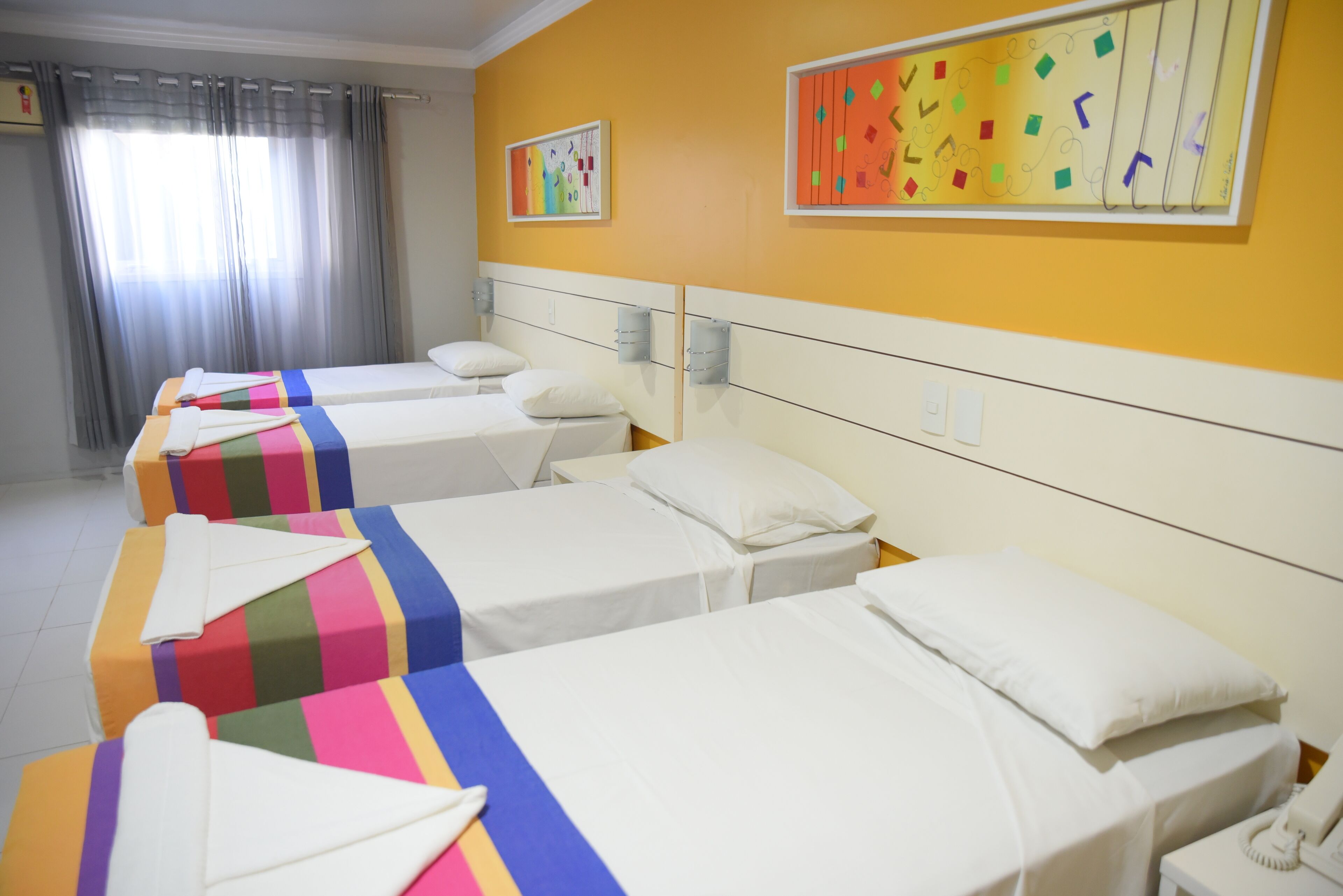 A hotel room with four beds, each with a colorful striped bedspread and white pillows. There are two wall-mounted light fixtures and a window with curtains.
