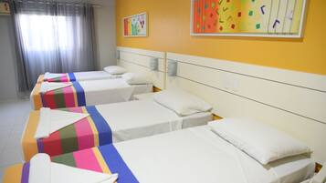A hotel room with four beds, each with a colorful striped bedspread and white pillows. There are two wall-mounted light fixtures and a window with curtains.