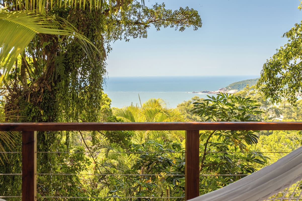 A hammock on a balcony overlooking a coastal landscape with dense greenery and a view of the sea.