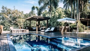 Un área de piscina con sillones de descanso, una zona de estar cubierta y palmeras al fondo.