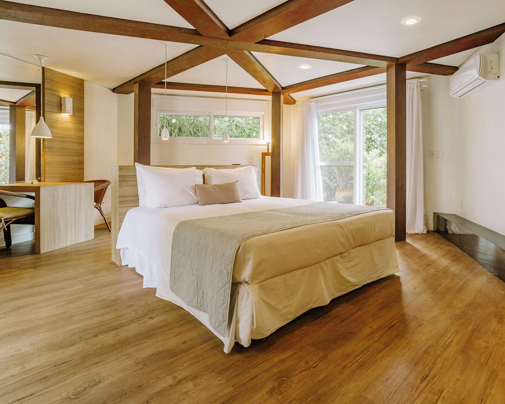 A bedroom with a large bed, wooden beams, a desk, and a window with a view of greenery.