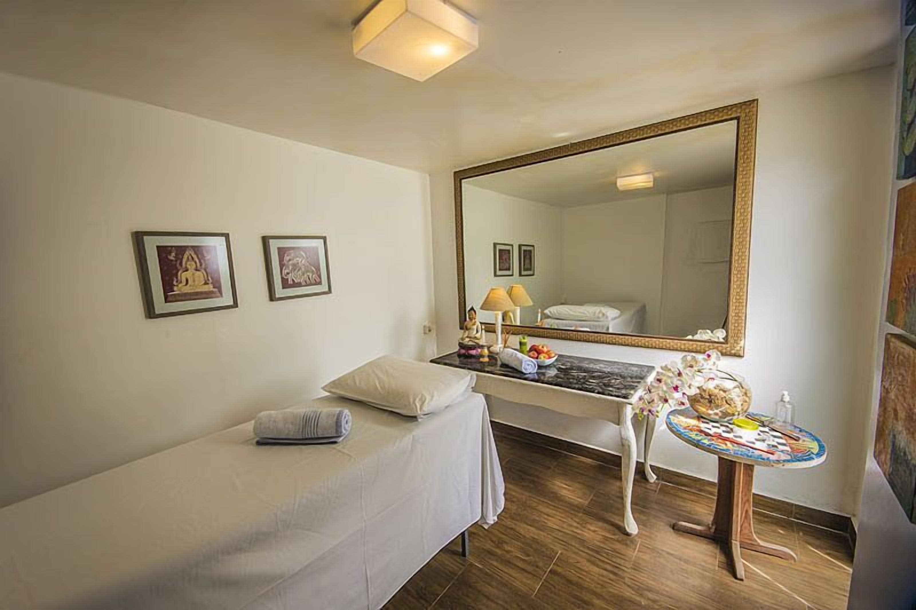 Habitación de hotel con cama, escritorio con espejo, pequeña mesa con lámpara y dos cuadros en la pared.