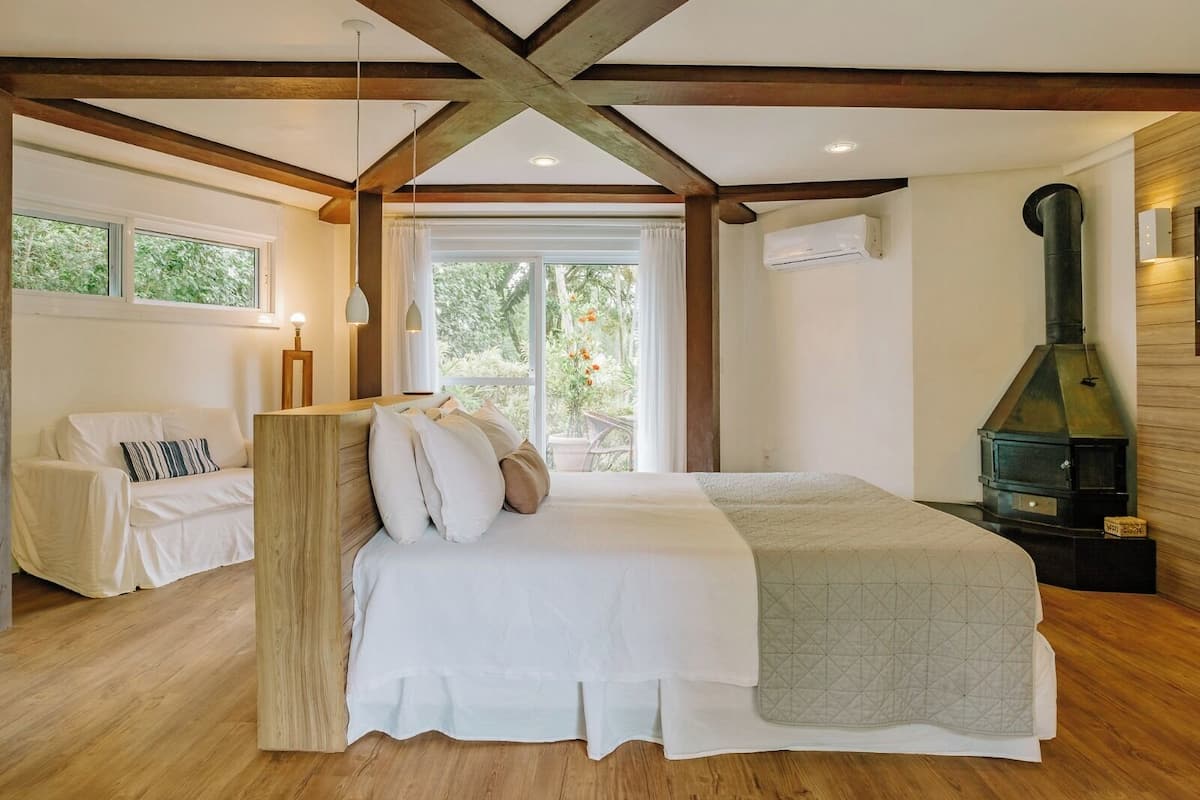 A bedroom with a large bed, wooden beams, a fireplace, and a view of greenery through the window.