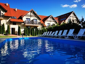 A swimming pool with lounge chairs, a building with red-tiled roofs, and a row of evergreen trees. - Hotel Santa Monica (Mikolajki)