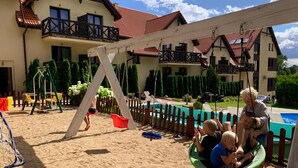A sandy outdoor area with a swing set, a pool, and a building with balconies. - Hotel Santa Monica (Mikolajki)
