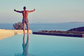 Un homme se tient au bord d’une piscine, d’où l’on aperçoit la mer et des collines au loin.