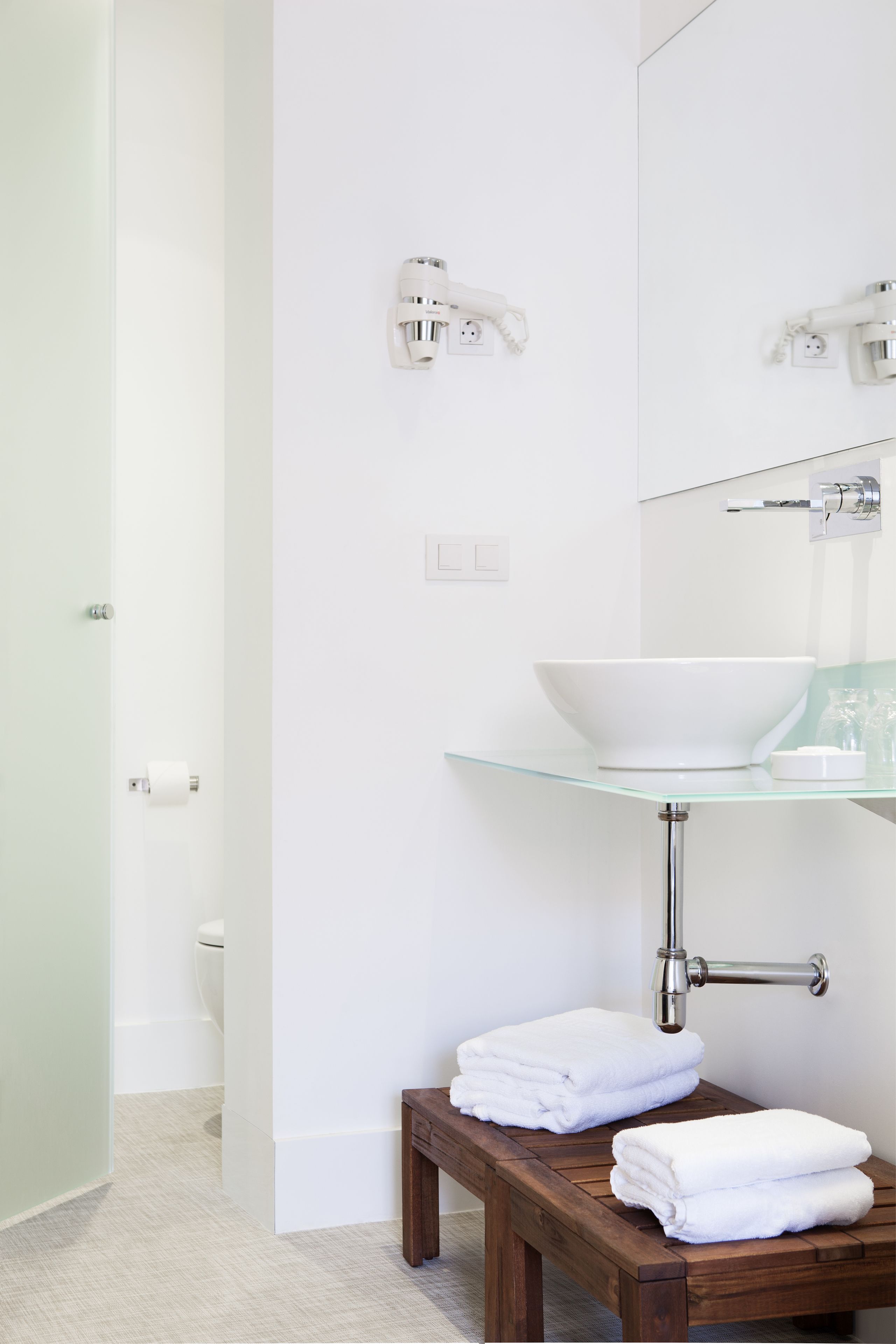 A modern bathroom with a glass sink, white fixtures, and a wooden bench with towels.