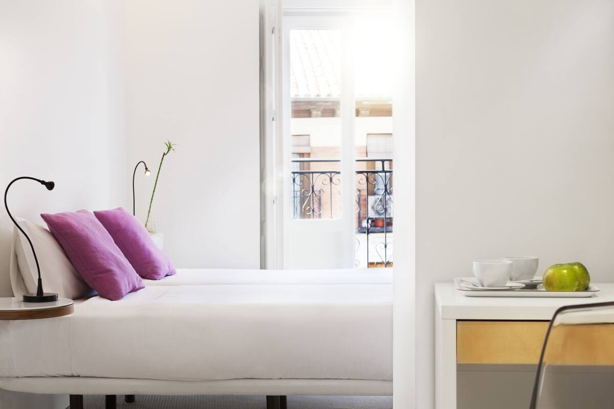 A minimalist room with a bed, a bedside table with a lamp, a small plant, and a view of a balcony with a railing.