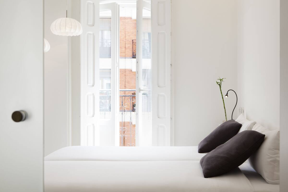 A bedroom with a white bed, two dark pillows, a hanging light fixture, and a view of a brick building through an open door.