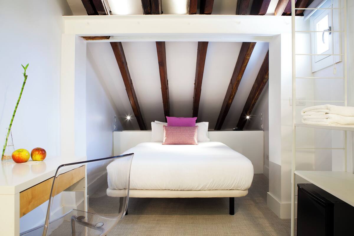 A modern bedroom with a sloped ceiling, a bed with white bedding, a pink pillow, and a bedside table with apples.