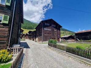 A traditional wooden building with a cobblestone street and a green hillside in the background. - Hotel Astoria (Obergoms)