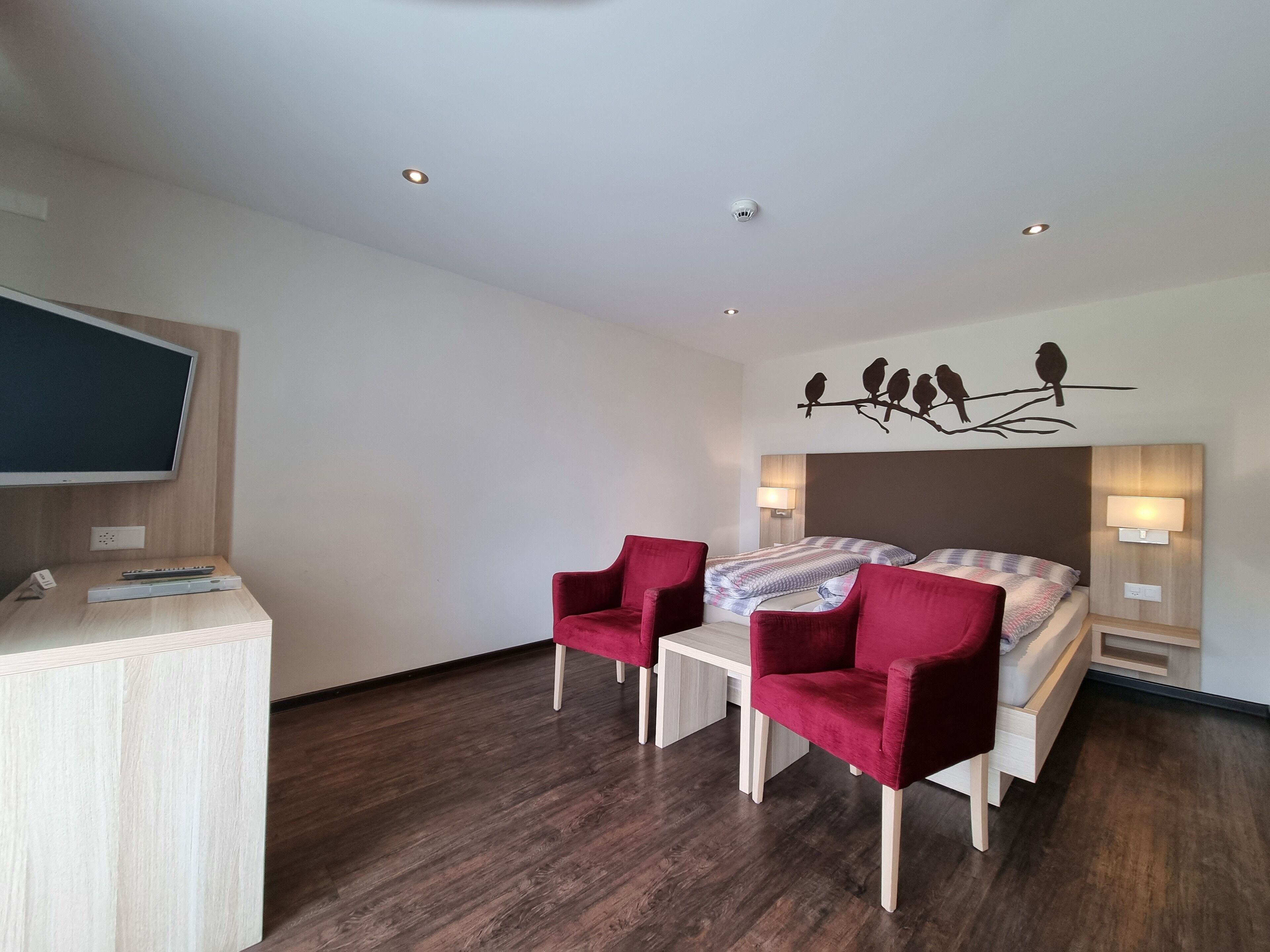 A modern hotel room with a bed, two red chairs, a TV, and a wall-mounted bird decoration.