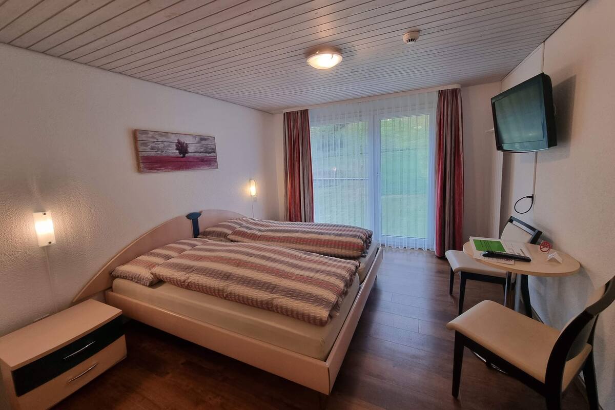 A bedroom with a bed, bedside tables, a desk with a chair, a TV, and a window with curtains.
