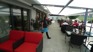 An outdoor seating area with red cushioned chairs and tables, surrounded by glass doors and windows. - Hotel Astoria (Obergoms)