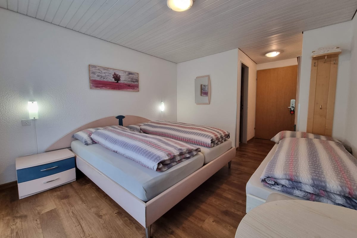 A room with two beds, a small bedside table with drawers, a wall-mounted light, and a door leading to another room.