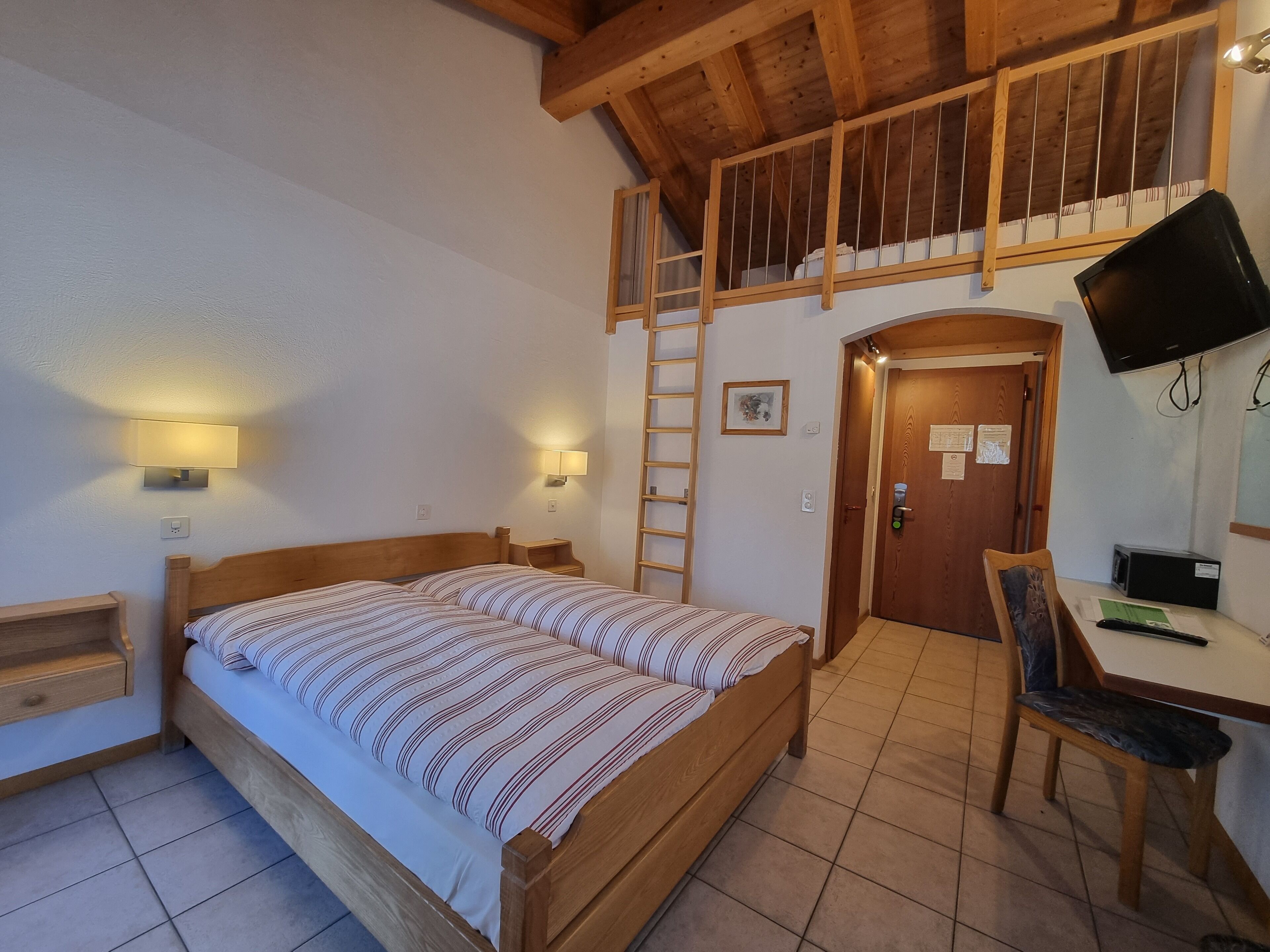 A bedroom with a wooden bed, a desk with a chair, a TV, and a ladder leading to a loft.