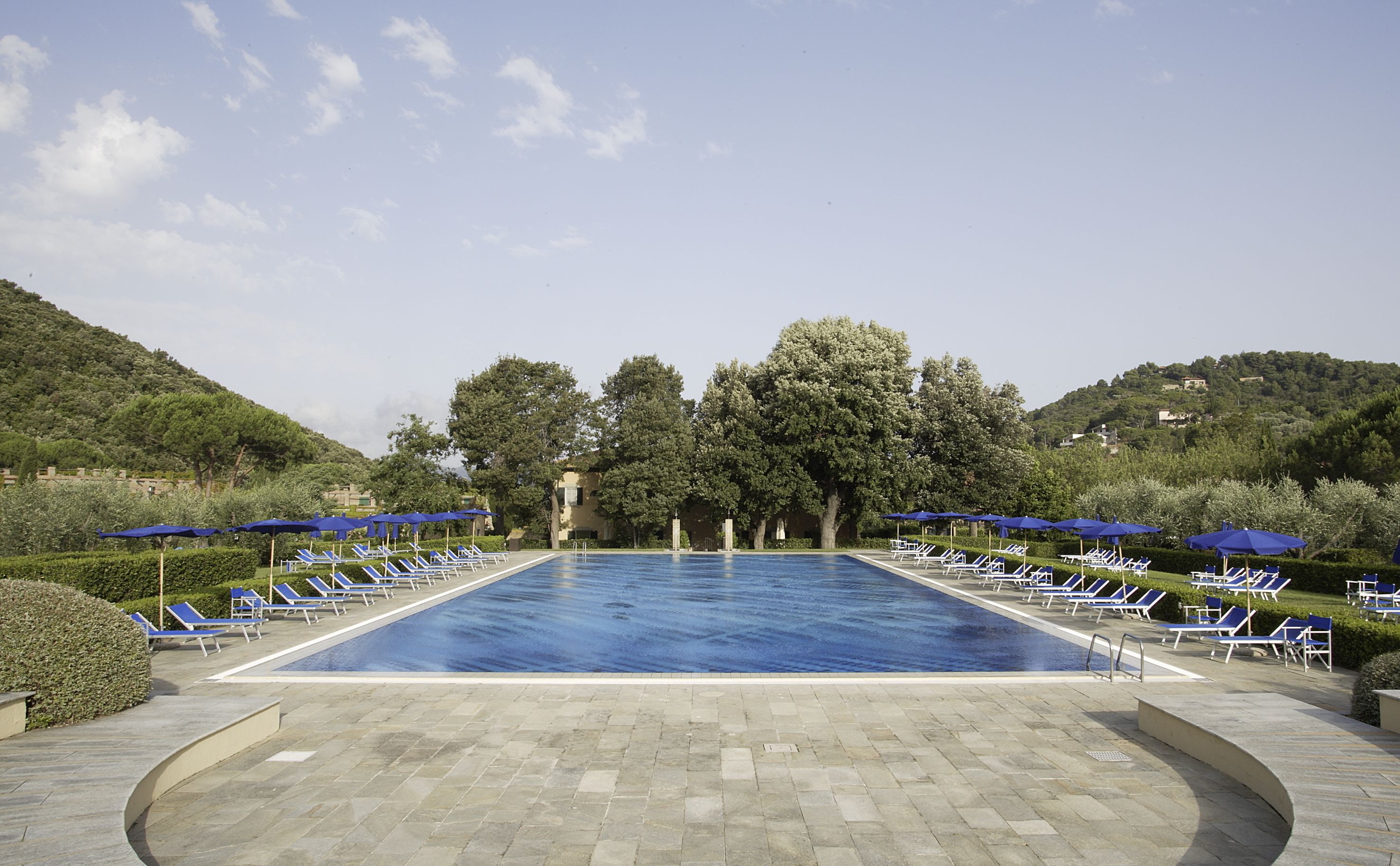 Ein großer rechteckiger Pool, umgeben von Liegestühlen und Sonnenschirmen, mit Bäumen und Hügeln im Hintergrund.