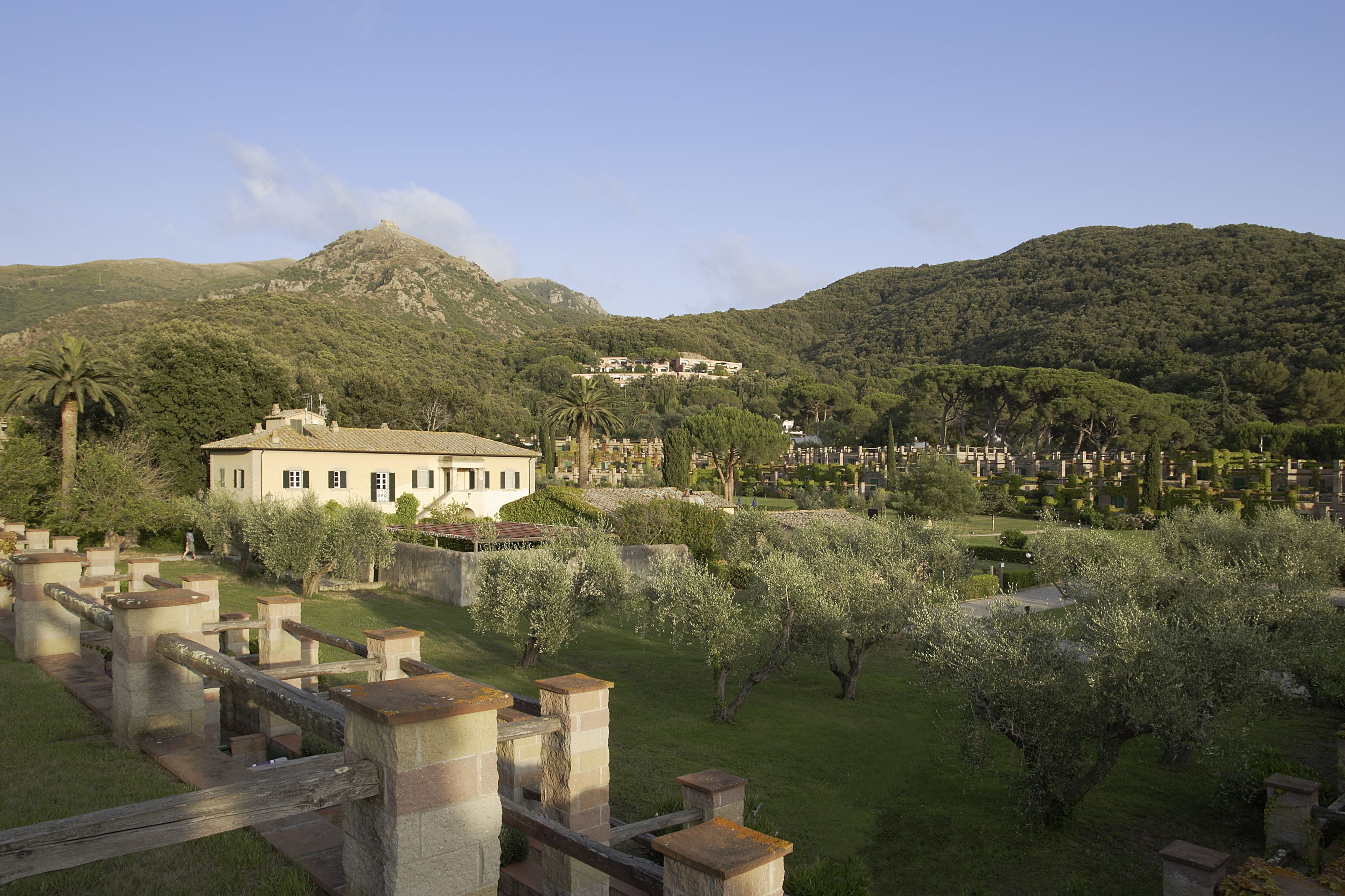 A large building with a white facade, surrounded by lush greenery and a mountainous backdrop.