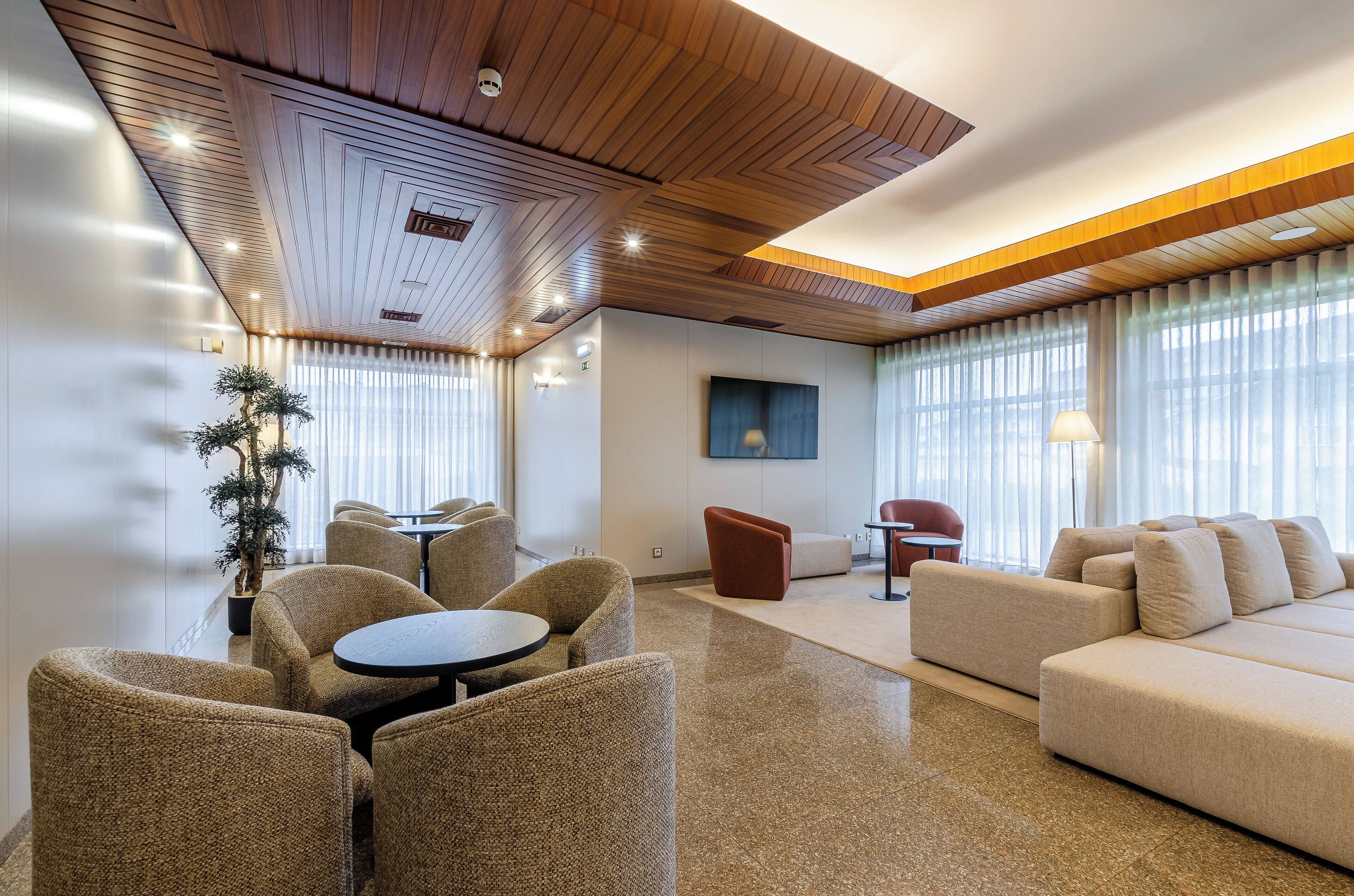 A modern hotel lobby with a wooden ceiling, a large sofa, a dining area with round tables, and a potted plant.