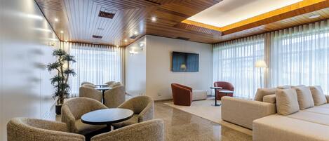 A modern hotel lobby with a wooden ceiling, a large sofa, a dining area with round tables, and a potted plant.
