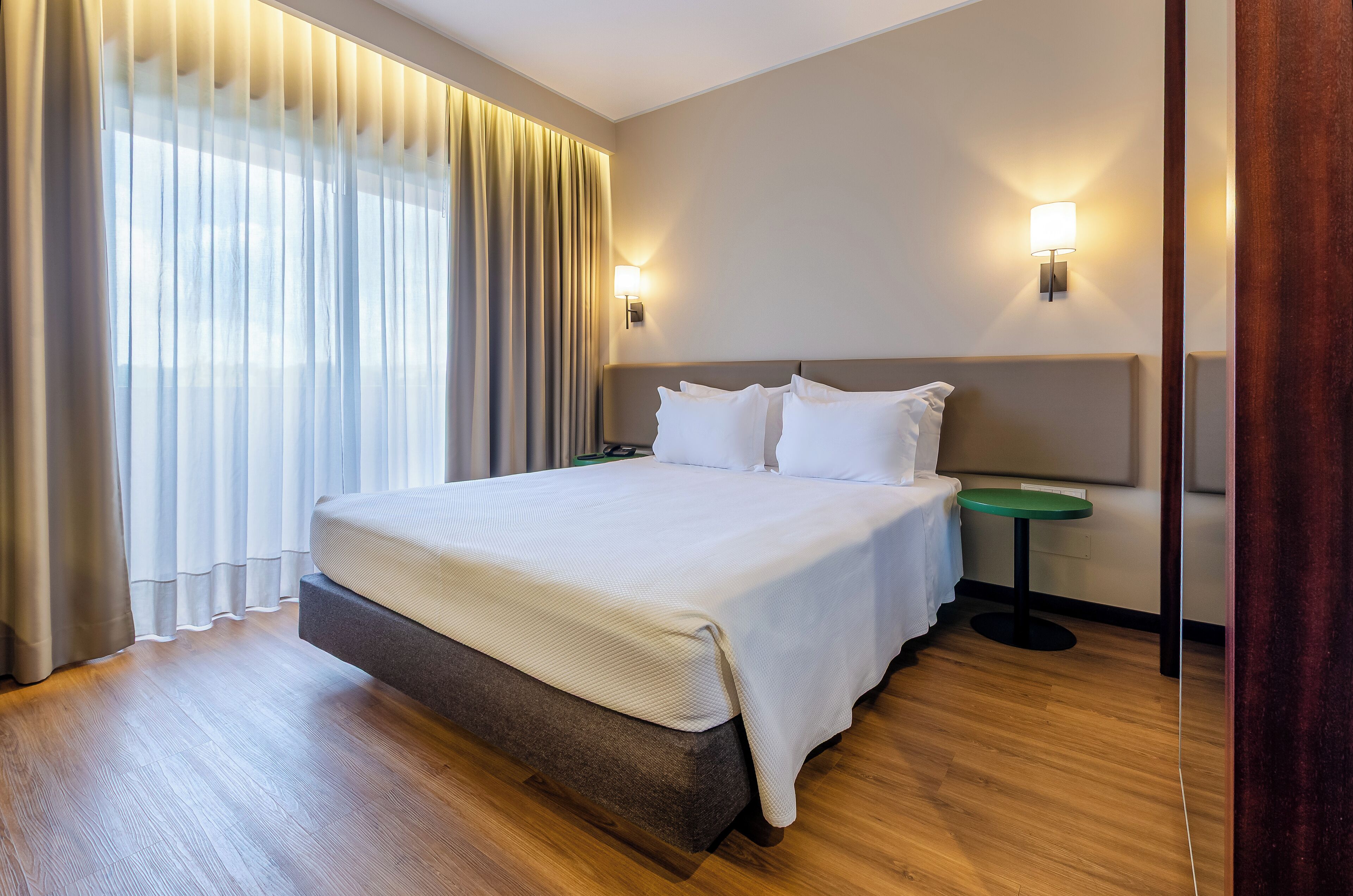 Habitación de hotel con una cama grande, dos mesitas de noche, una mesita auxiliar verde y una ventana con cortinas.