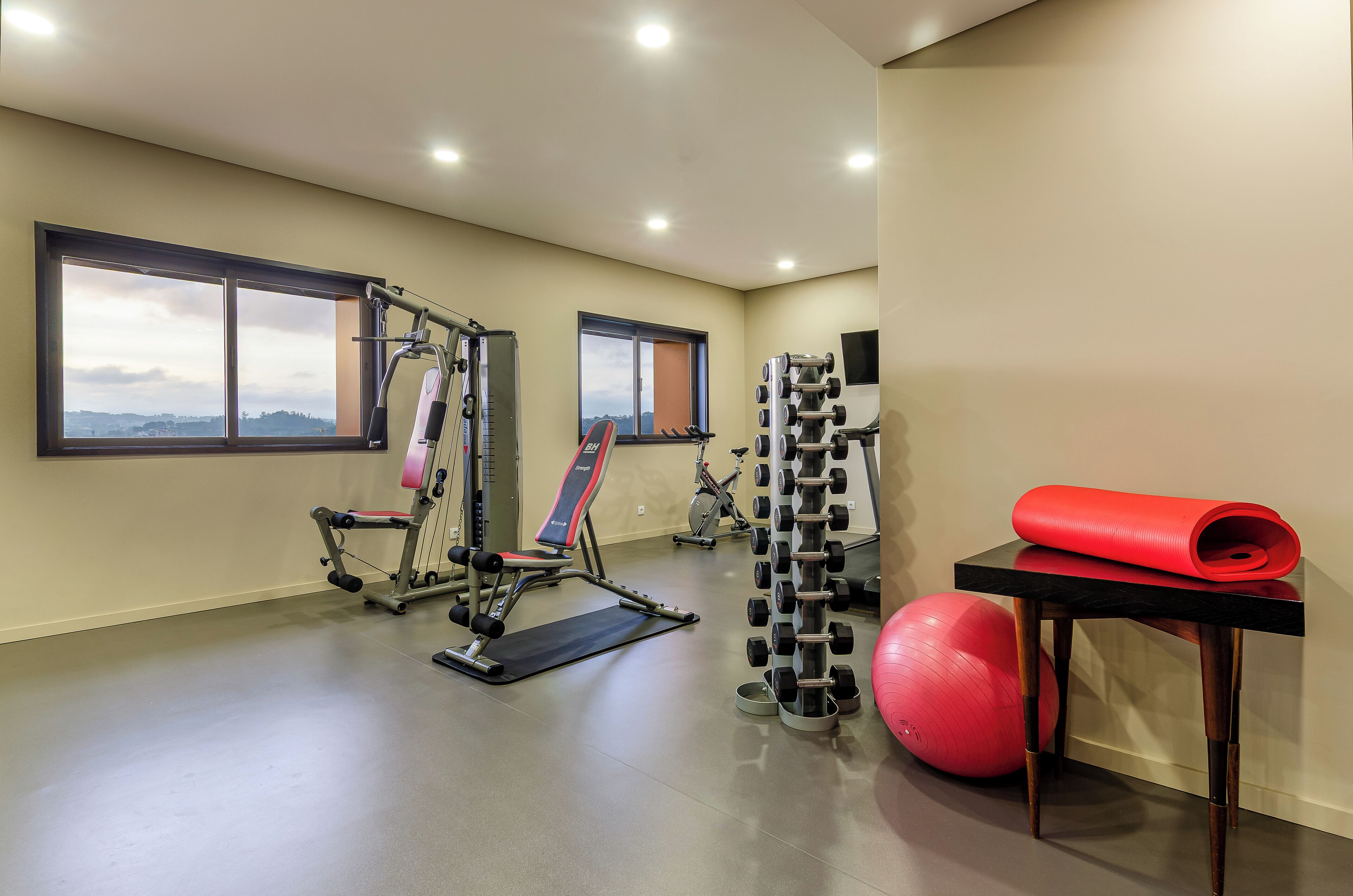 Une salle de sport équipée de matériel d’exercice, avec des fenêtres et un tapis de yoga rouge posé sur une table.