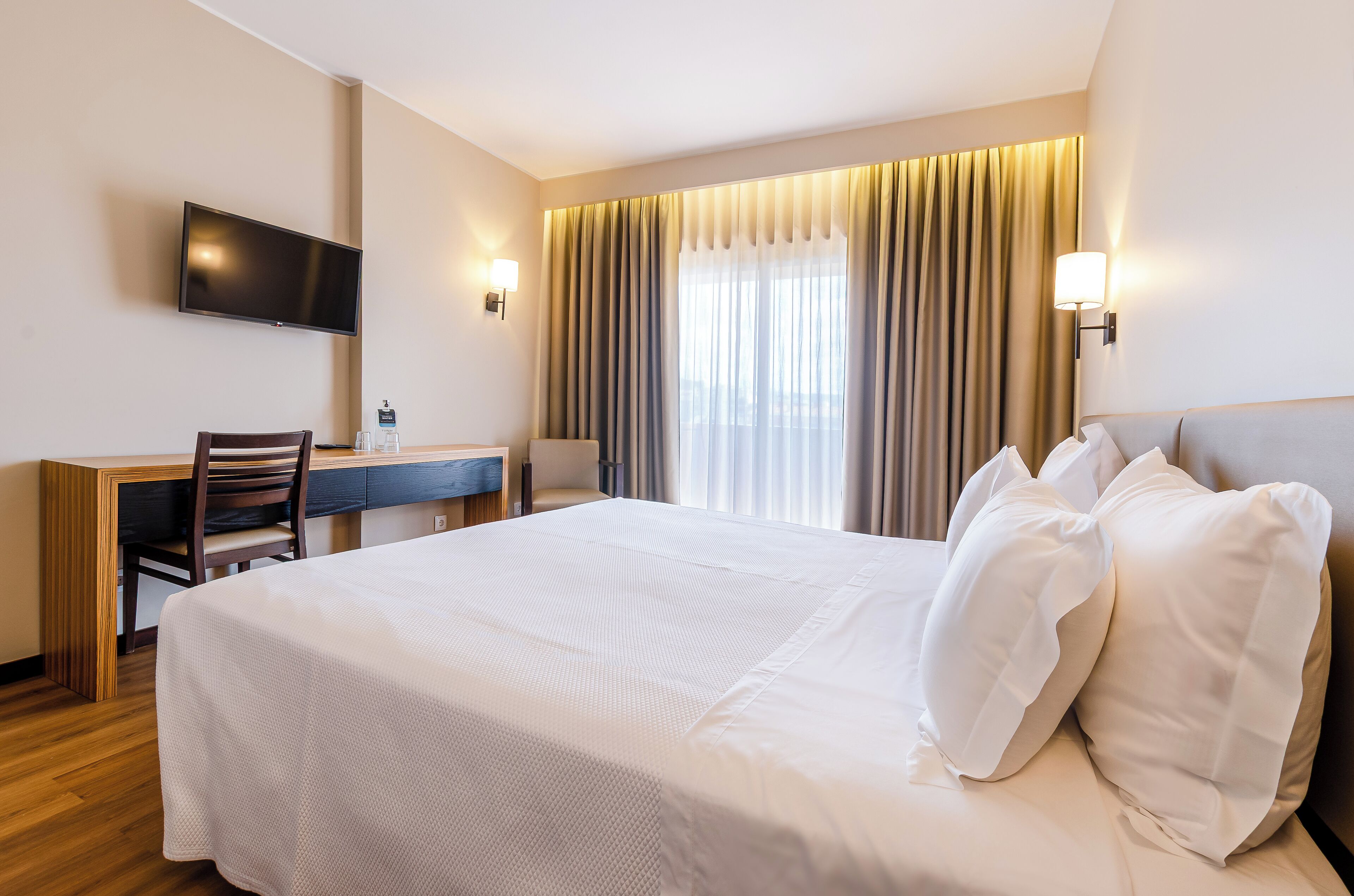 A hotel room with a large bed, a desk with a chair, a television mounted on the wall, and a window with curtains.