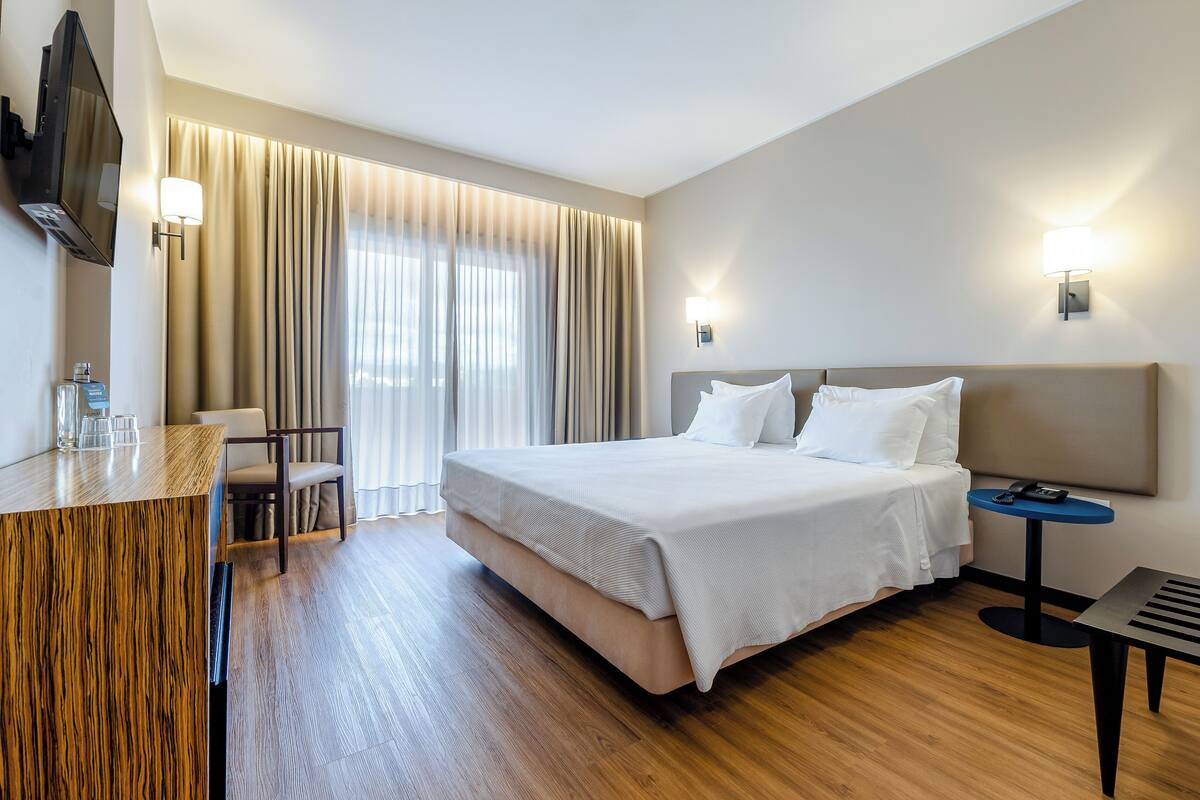 Une chambre d’hôtel comprenant un lit, un bureau avec une chaise, un téléviseur fixé au mur et une fenêtre avec des rideaux.